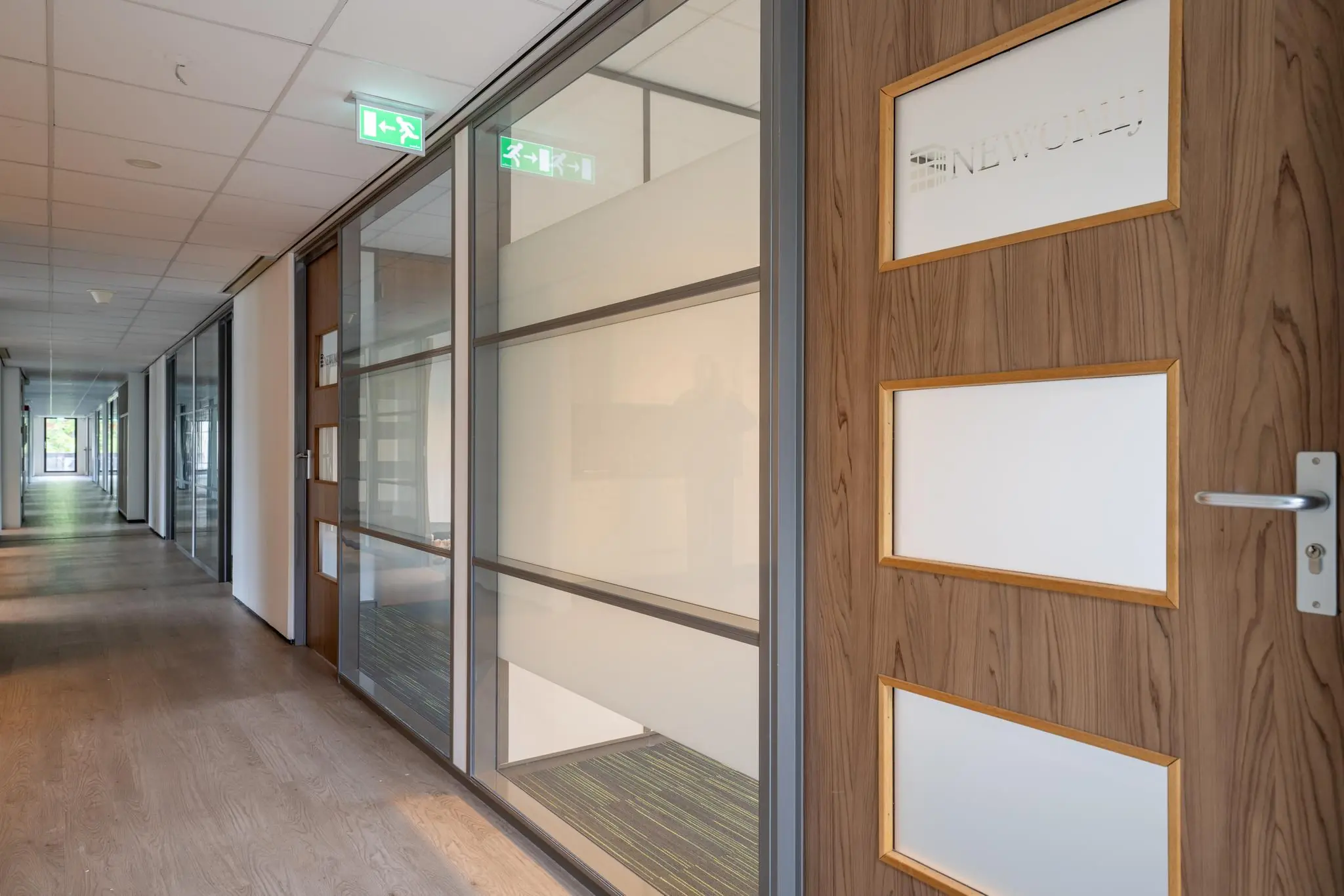 Moderne kantoorhal met houten deur van kantoorruimte 'NEWOMIJ' en glazen wanden langs een lange gang.